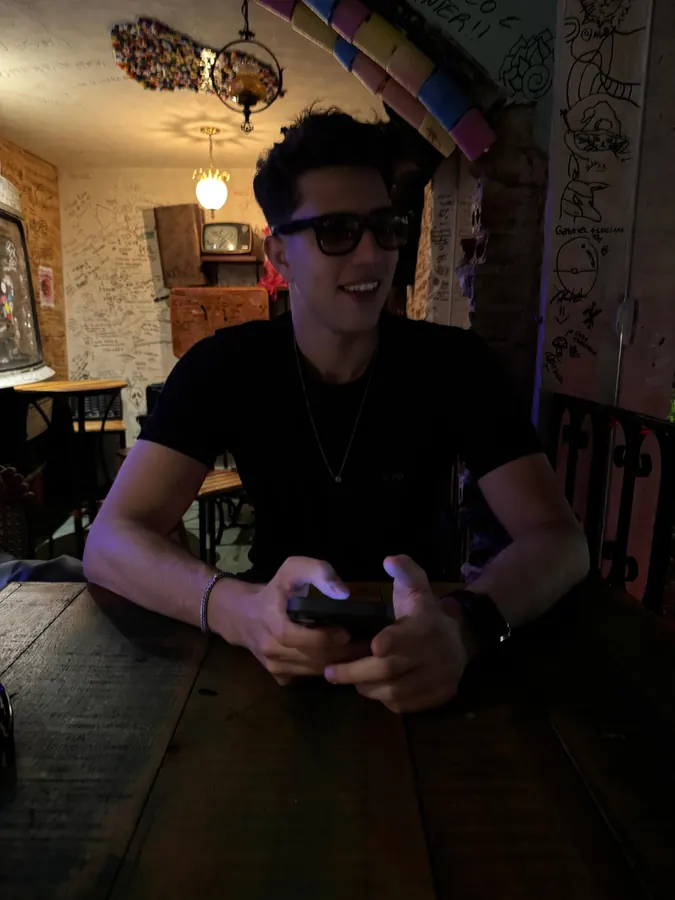 Matheus Barros em um bar com óculos escuros, sorrindo e segurando o celular